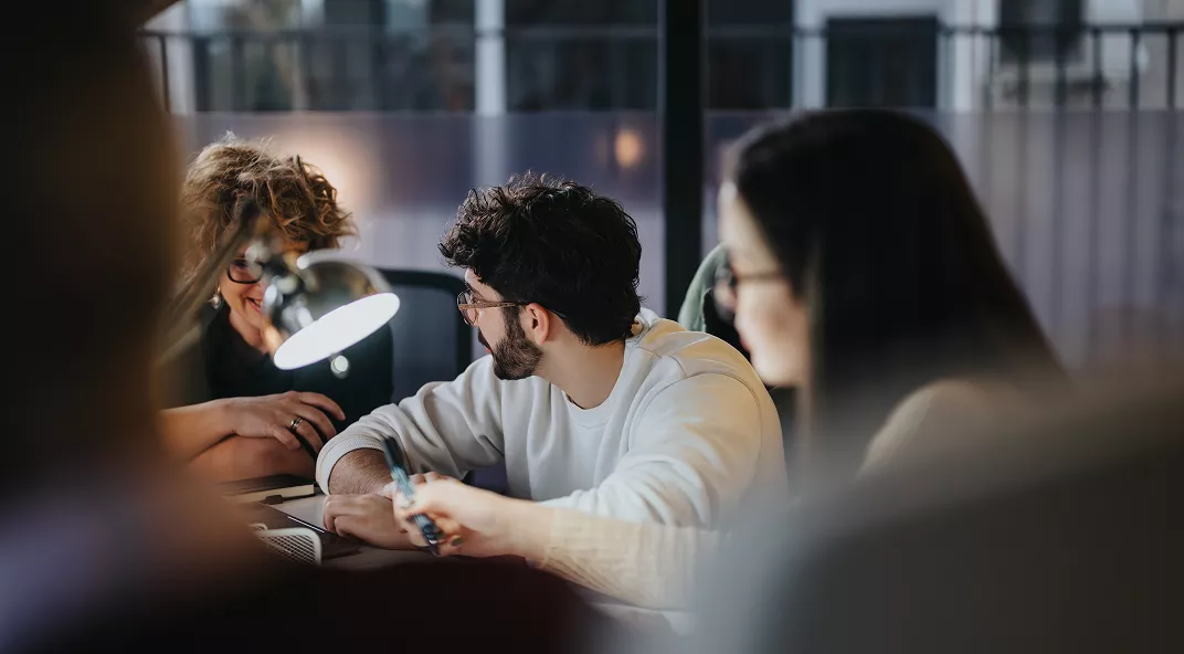 A group of young adults are gathered around a table in a contemporary office environment, engaged in discussion and teamwork. The workspace features a desk lamp and large windows, creating a bright and focused atmosphere. The individuals appear to be sharing ideas, with one person holding a pen and others listening attentively. The overall mood is collaborative and productive, with a neutral color palette and casual attire.