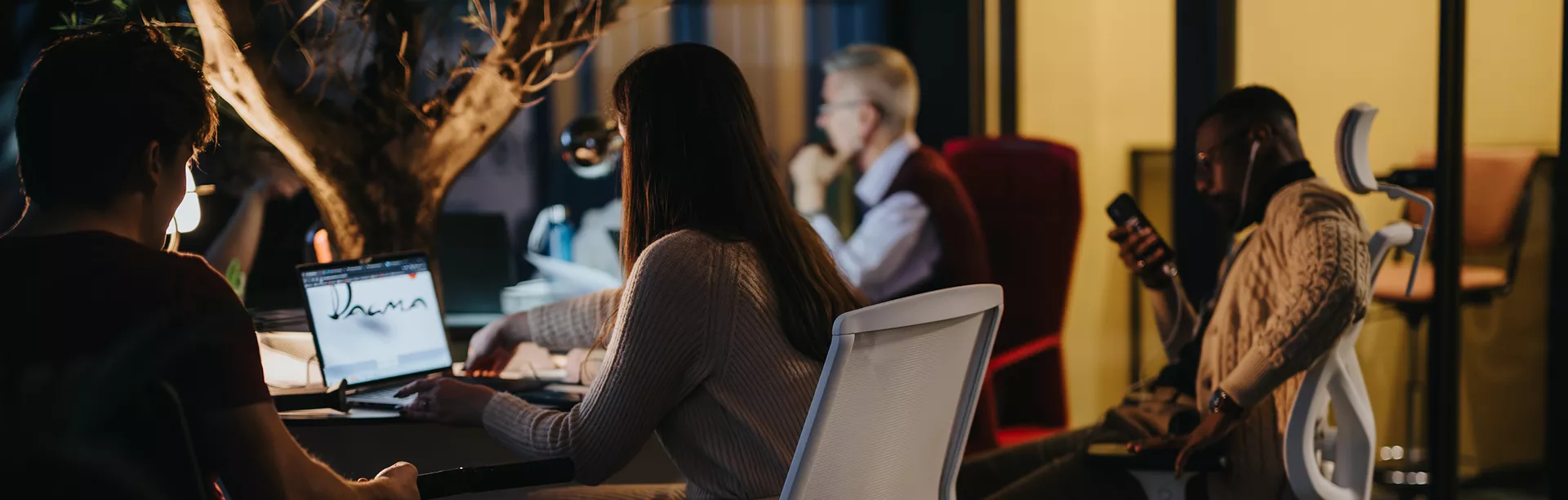 A group of adults work together in a contemporary office environment, illuminated by warm lighting. One person is using a laptop displaying the word 'Dreams,' while another checks their phone nearby. The workspace features a large indoor plant and glass partitions, creating a stylish and productive atmosphere.