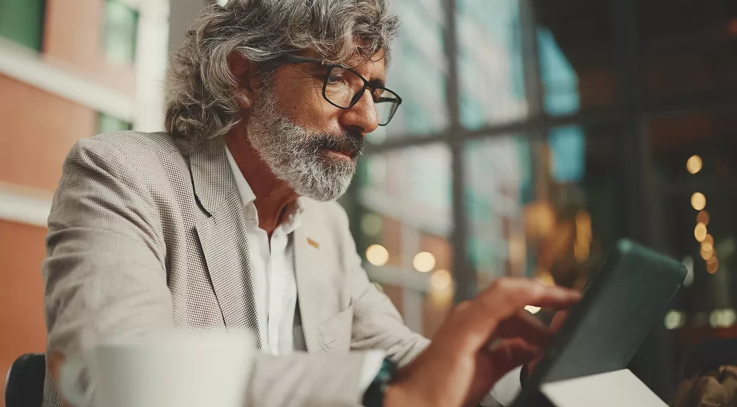 A mature man with gray hair and glasses is seated indoors, interacting with a tablet device. He is dressed in a light-colored suit jacket and white shirt, suggesting a professional or business environment. The background features large windows and blurred architectural elements, indicating a contemporary office or workspace. The overall mood is focused and professional, with warm, natural lighting.