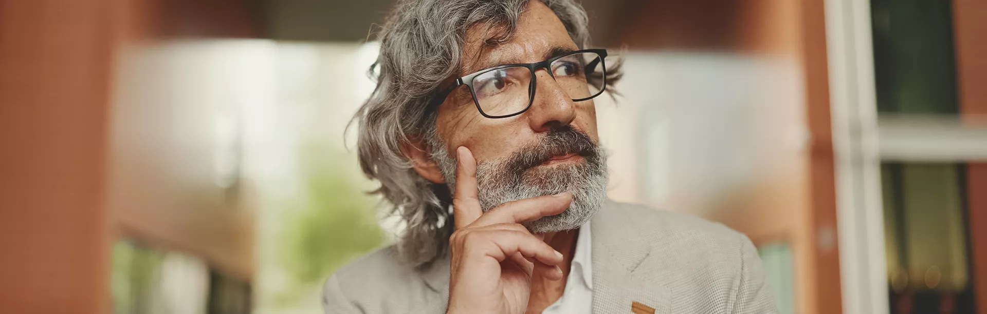 An older man with gray hair and a beard is seen wearing glasses and a light-colored blazer. He is outdoors in an urban setting, resting his chin on his hand in a contemplative pose. The background features blurred modern architecture and soft natural light, creating a reflective and intellectual mood.