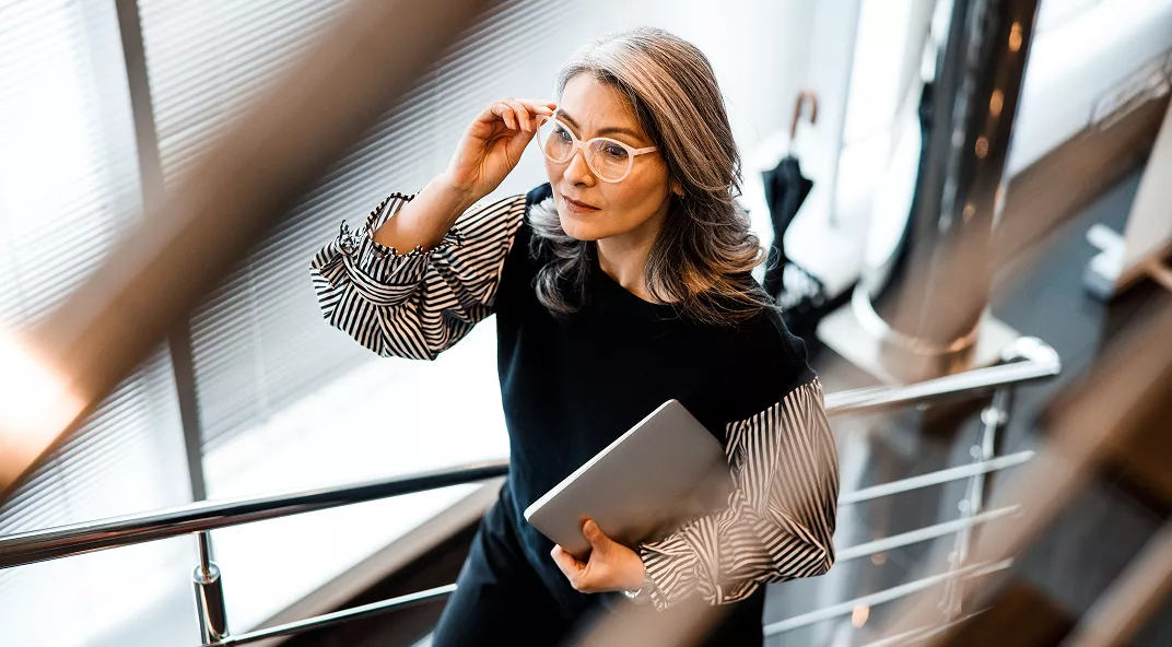 A mature woman with gray hair stands on a modern staircase, holding a tablet in one hand. She is dressed in a stylish black and white outfit and appears to be adjusting her glasses while looking thoughtfully into the distance. The setting is a bright, contemporary office space with large windows and metallic accents.