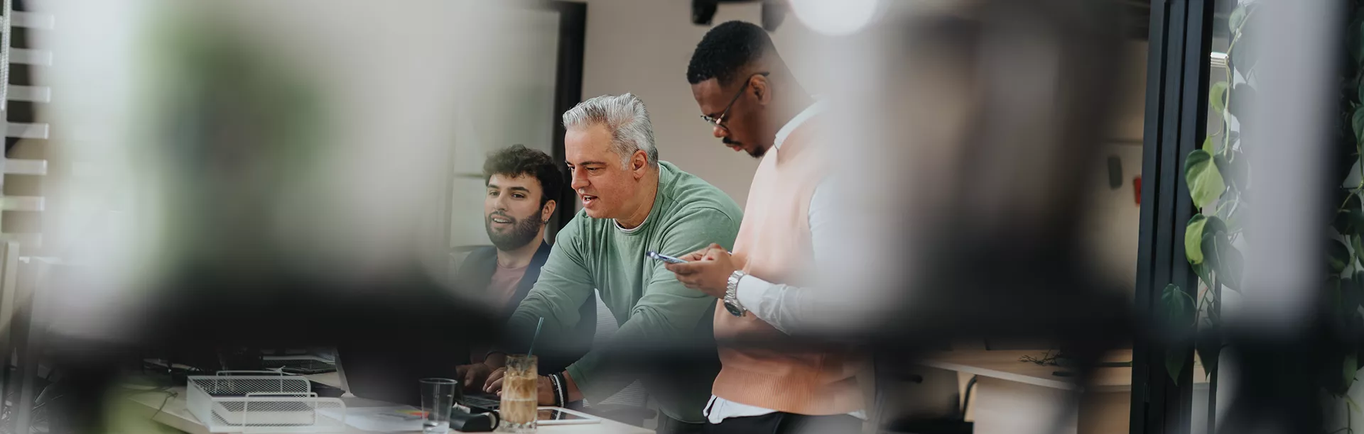 A group of three people are gathered around a desk, engaged in discussion and teamwork. The setting is a contemporary office with natural light and greenery visible in the background. The individuals appear to be of different ages and backgrounds, contributing to a collaborative atmosphere. The image is shot through a glass partition, adding a candid and dynamic visual style.