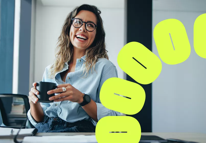 A woman with wavy hair sits casually at a desk in a modern office environment, holding a black coffee mug. She is dressed in a light blue button-up shirt and wears a black wristwatch. The setting features contemporary decor with bright yellow graphic elements in the foreground, adding a creative touch to the scene.