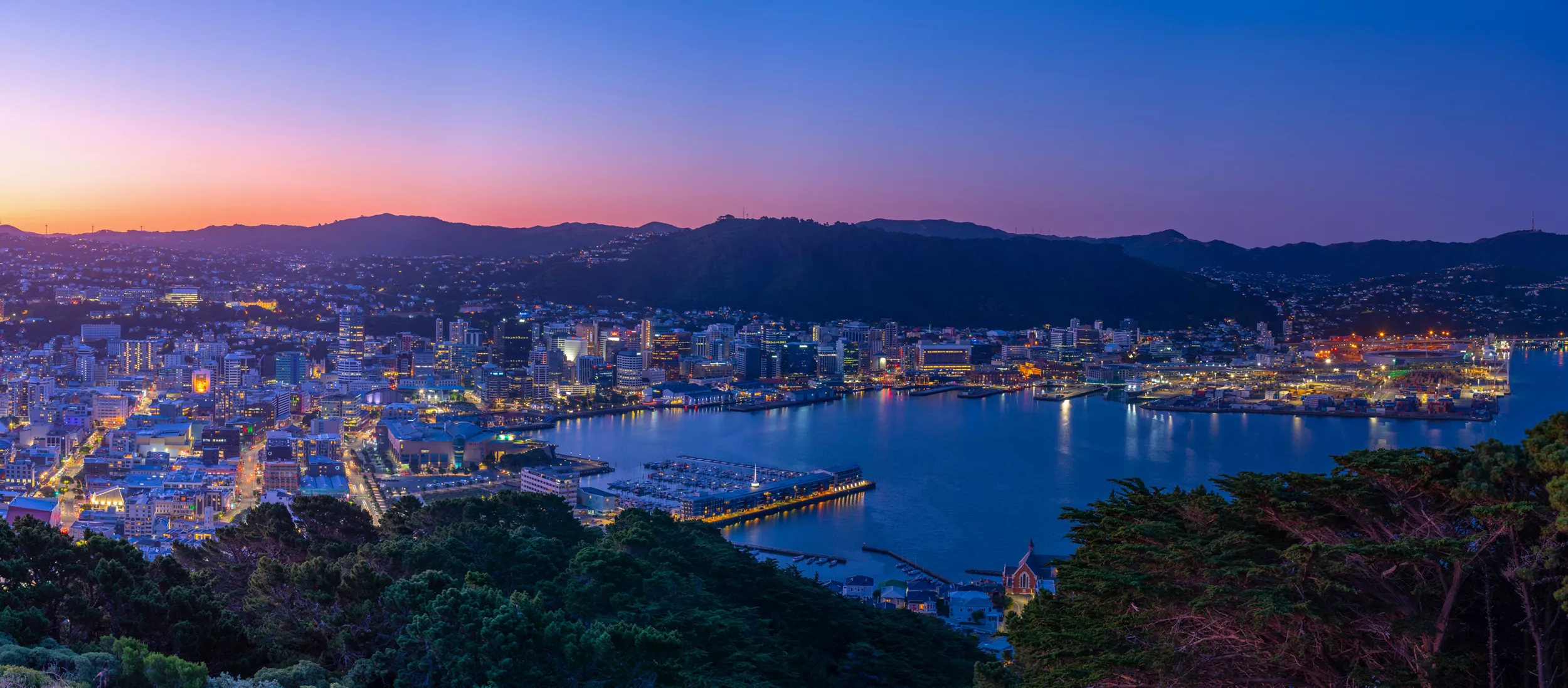 Panoramic view of a coastal city and harbor captured at twilight. The scene shows illuminated buildings and waterfront lights reflecting on calm water. Hills and mountains form a dark silhouette against a vibrant gradient sky transitioning from warm sunset tones to deep blue. Foreground trees frame the urban landscape, emphasizing the contrast between nature and city.