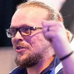 A bearded man with glasses is captured mid-speech, gesturing with his hand during a live presentation. He has long hair tied back and appears to be speaking passionately. The background is softly lit with a bluish tone, suggesting a stage or conference environment. The close-up framing emphasizes his facial expression and animated gesture.