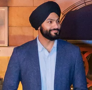 A bearded man wearing a dark turban and blue blazer stands indoors, looking to the side with a slight smile. He is dressed in a light blue shirt, giving a polished and professional appearance. The warm-toned background features architectural details and soft lighting, suggesting an event or social gathering. The overall scene conveys confidence and a relaxed, formal atmosphere.