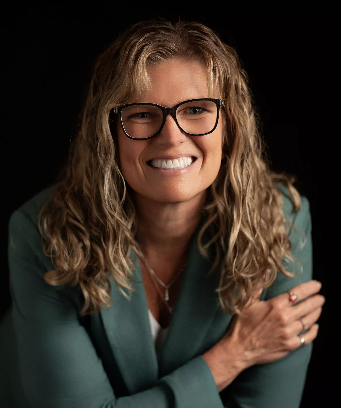 Portrait of a smiling woman with wavy blonde hair wearing dark-framed eyeglasses. She is dressed in a teal blazer over a light top, posed with arms crossed against a dark background. The image has a warm, professional feel suitable for business or personal branding. Soft lighting highlights her face and creates a polished, studio-style look.