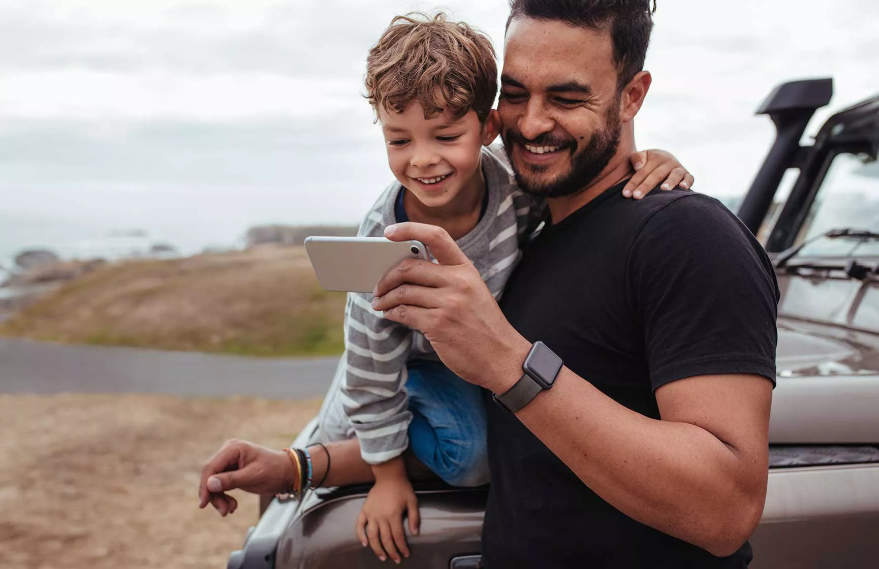 An adult and a child are standing together by a parked vehicle in an outdoor setting, possibly near a coastline or open field. The adult is holding a smartphone, and both appear to be looking at the screen, suggesting a shared moment or activity. The adult is wearing a smartwatch and a black t-shirt, while the child is dressed casually. The background features natural scenery with muted earth tones.