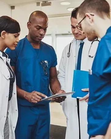 Two doctors in white coats and two in blue scrubs standing in a circle and leaning over a tablet.