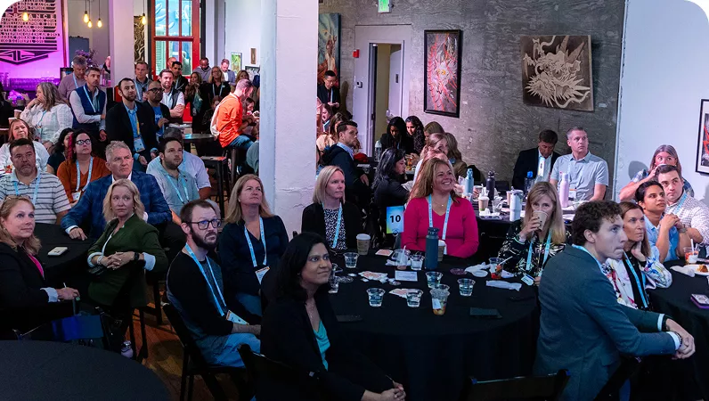 A professional gathering featuring a diverse audience seated at round tables in a modern venue. The setting includes artwork on the walls and visible table numbers, such as '10'. Attendees are wearing business attire and conference lanyards, suggesting a formal networking or presentation event.