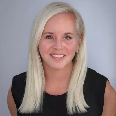 Portrait of a smiling woman with long straight blonde hair, looking directly at the camera. She is wearing a sleeveless black top against a neutral light gray background. The image is well-lit, highlighting her facial features and friendly expression. This appears to be a professional headshot suitable for corporate or business use.