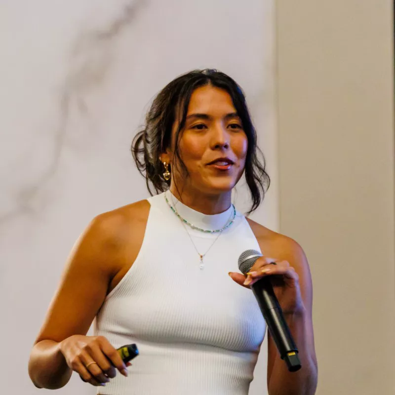 A young woman stands indoors holding a handheld microphone while speaking. She wears a sleeveless white top and layered necklaces, with her hair tied back. The background is softly lit with neutral tones, suggesting a presentation or event setting. Her expressive posture and slight smile convey an engaging, confident mood.