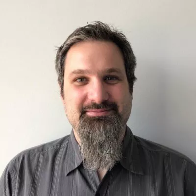 Portrait of an adult man with a long gray beard standing against a plain light-colored wall. He is wearing a dark button-up shirt and looking directly at the camera with a slight smile. The lighting is soft and even, creating a neutral, professional appearance. The simple background keeps the focus on the subject’s face and expression.