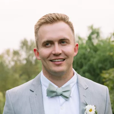 A young adult man is dressed in a light gray suit with a matching bow tie, standing outdoors. He is smiling and looking slightly away from the camera, with soft natural light on his face. Green trees and foliage create a blurred background, suggesting a garden or park setting. A small white flower is pinned to his lapel, adding a subtle decorative detail.
