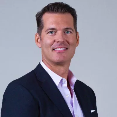 Portrait of a smiling adult man wearing a dark blazer over a light pink dress shirt. He is posed against a plain light gray background, looking slightly to the side of the camera. The image has a clean, professional look suitable for corporate or business contexts. Lighting is soft and even, highlighting his face and formal attire.