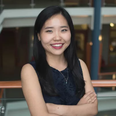 A young woman stands indoors with her arms crossed, smiling warmly at the camera. She is dressed in a dark sleeveless top and wears a delicate necklace, suggesting a professional or business-casual setting. The background shows an office or institutional interior with railings, glass, and architectural details softly out of focus. The overall mood is confident, approachable, and friendly.