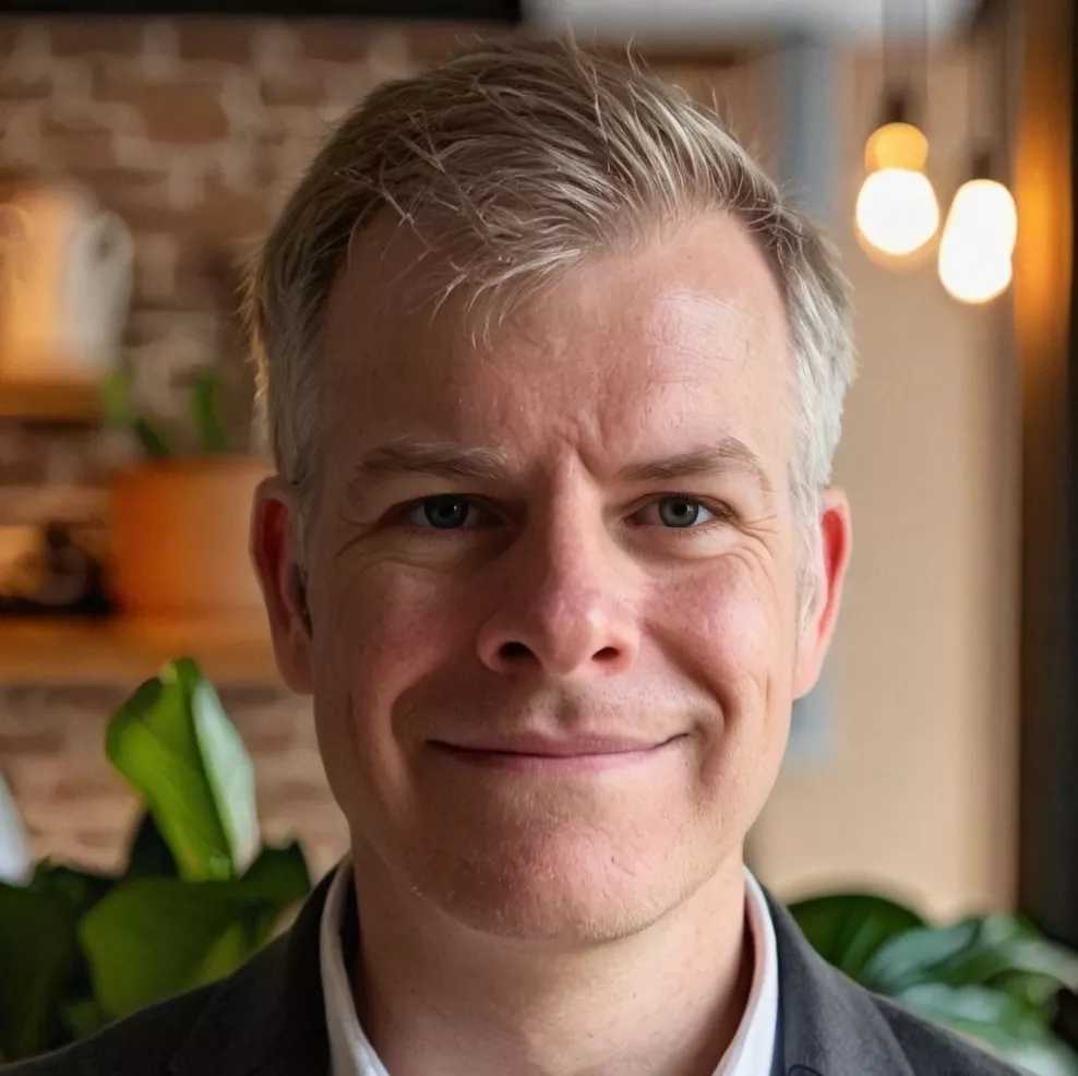 Close-up portrait of a smiling adult man with short, light-colored hair. He is indoors in a contemporary space with warm lighting and a softly blurred background. Brick and wooden elements, plants, and hanging lights create a cozy, modern atmosphere. The image conveys a friendly, approachable, and professional mood.