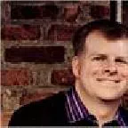 Portrait of a smiling adult man standing in front of a rustic brick wall. He is wearing a dark jacket over a collared shirt with visible stripes. The image is closely cropped, focusing on his face and upper torso. The warm tones of the bricks create a casual, friendly atmosphere.