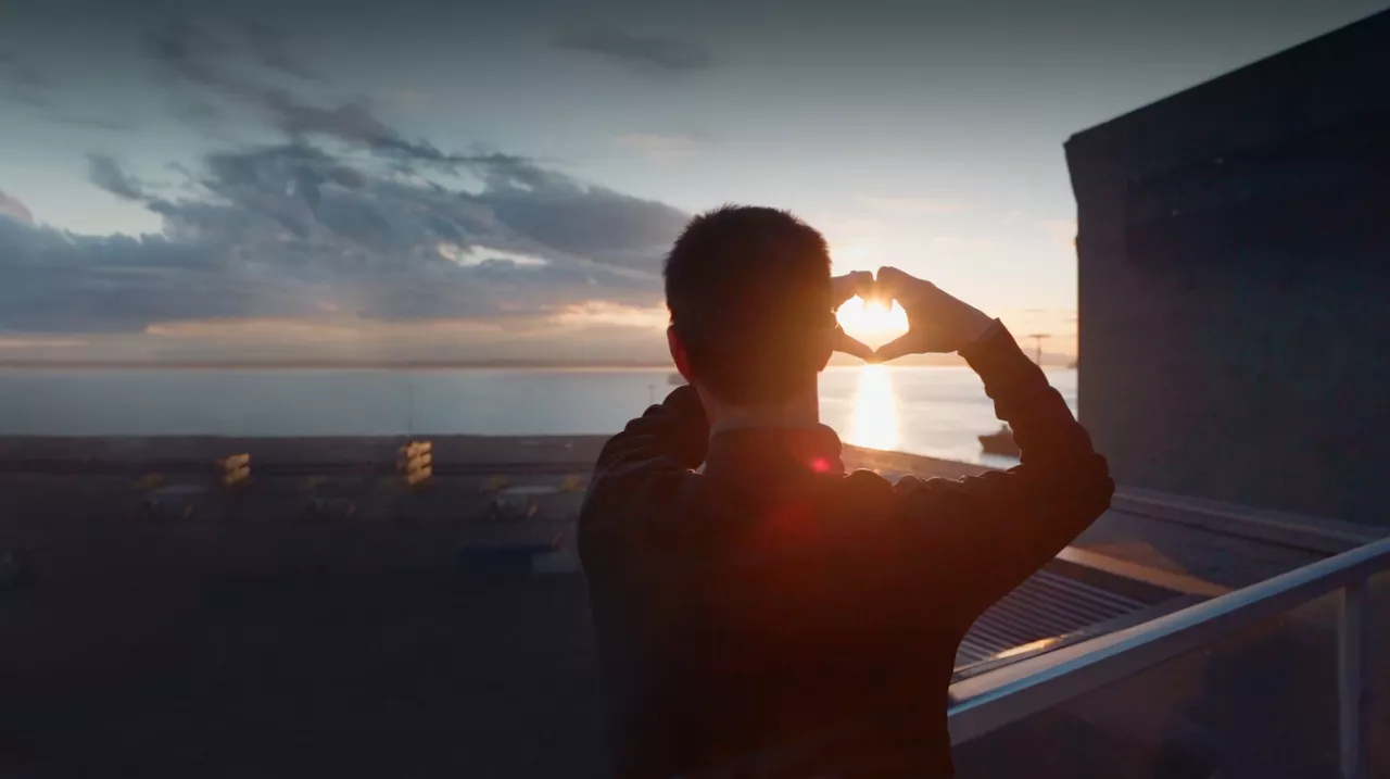 Man on balcony holding hands up in the shape of a heart around the setting sun