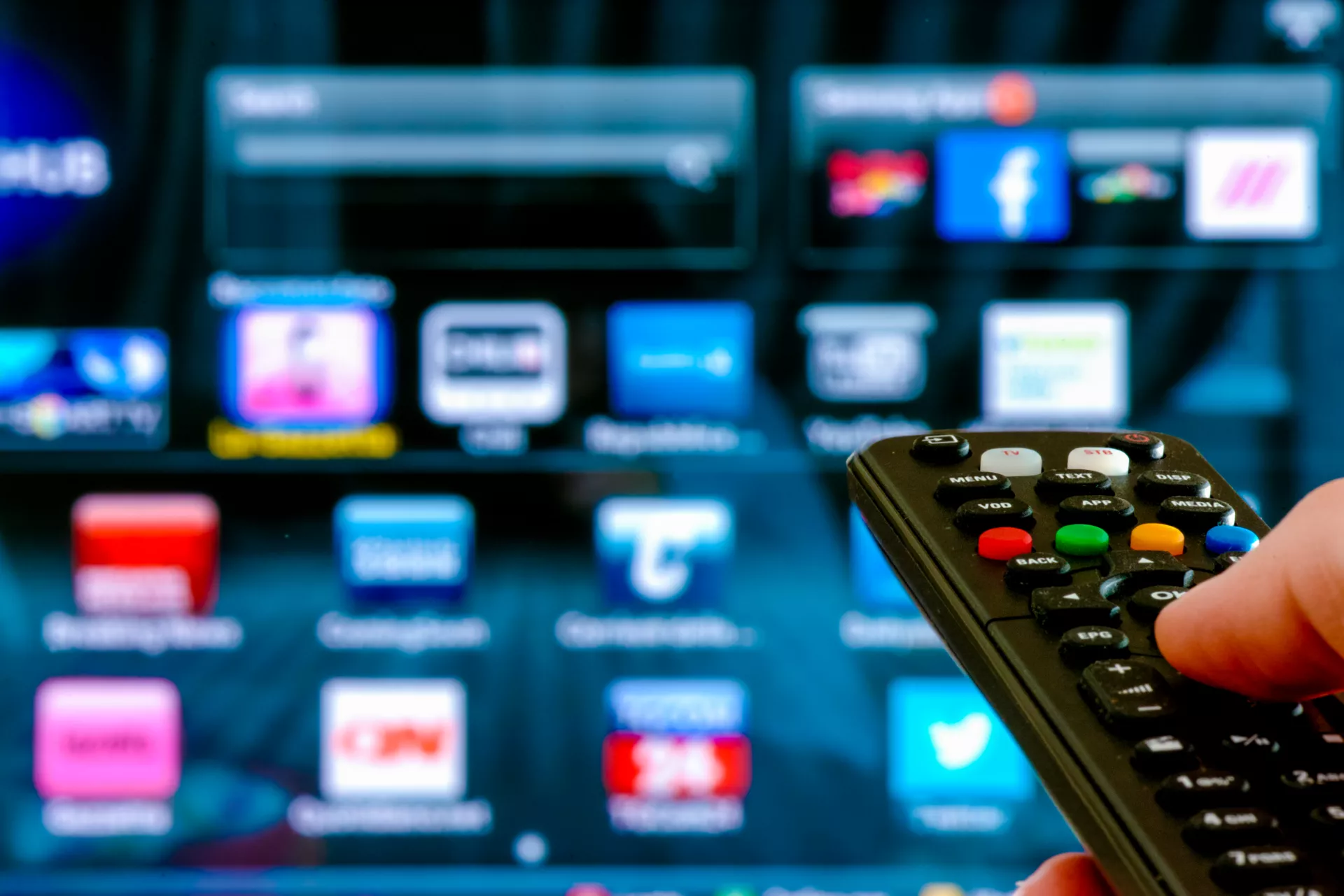 A person holds a television remote control pointed at a smart TV screen. The TV interface displays colorful app icons and menus in a grid layout. The scene suggests browsing or selecting streaming and online services. Bright, saturated colors and a shallow depth of field emphasize the remote in the foreground and the digital interface in the background.