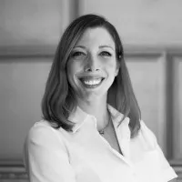 Portrait of a smiling woman wearing a white button-up shirt. She is standing indoors against a softly lit, textured wall. The image is in black and white, emphasizing contrast and facial expression. The overall composition conveys professionalism, approachability, and confidence.