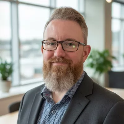 Portrait of a bearded man wearing glasses and a blazer, standing in a bright contemporary office space. Large windows in the background let in natural light and reveal an out-of-focus urban view. Indoor plants and light-colored furniture contribute to a clean, modern atmosphere. The subject appears approachable and confident, suitable for corporate or professional contexts.