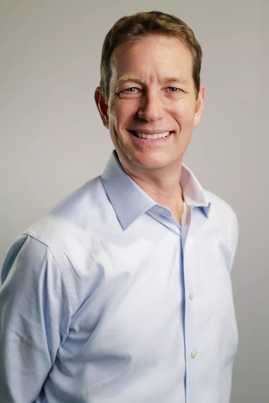 A middle-aged man smiles warmly at the camera in a studio setting. He is wearing a light blue button-up shirt and is posed slightly at an angle. The background is plain and neutral, keeping the focus on his friendly expression. The overall image conveys approachability and professionalism.