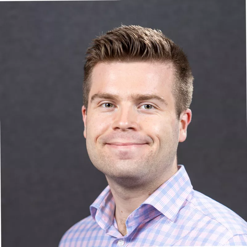 A smiling man is posed for a professional headshot against a dark gray studio background. He wears a light pink and blue checkered button-down shirt with the collar open. His hair is neatly styled, and he looks directly at the camera with a friendly expression. The simple backdrop and soft lighting emphasize his face and upper body.