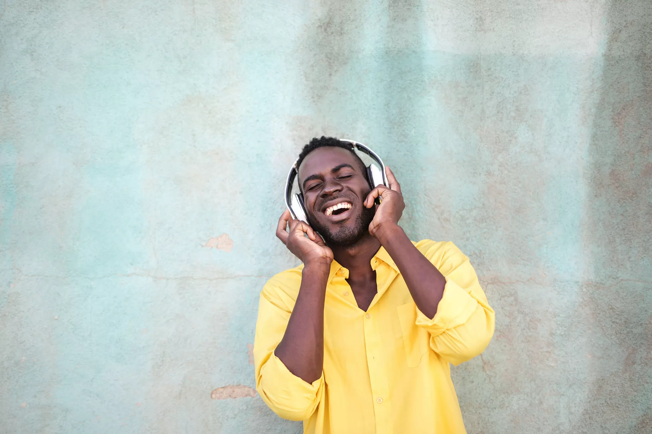 A young man in a bright yellow shirt enjoys music through over-ear headphones. He stands against a textured, pastel-colored wall, smiling with his eyes closed. His hands hold the headphones close, suggesting he is immersed in the sound. The overall scene conveys relaxation, happiness, and carefree enjoyment.