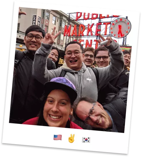 A diverse group of friends poses for a cheerful selfie in front of a brightly lit public market sign. The scene appears to be in an urban outdoor setting with buildings and storefronts in the background. Several people flash peace signs and smile enthusiastically toward the camera. The overall mood is fun and energetic, highlighted by emojis of a United States flag, a peace hand sign, and a South Korean flag beneath the photo.