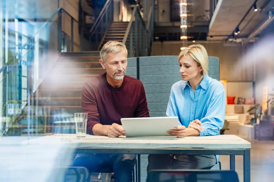 Two adults are seated at a table in a contemporary office environment, engaged in discussion while looking at a tablet device. The setting features industrial design elements, including exposed concrete and glass, with a staircase in the background. A glass of water is visible on the table, adding to the casual and collaborative atmosphere. The lighting is bright and natural, contributing to a professional yet relaxed mood.