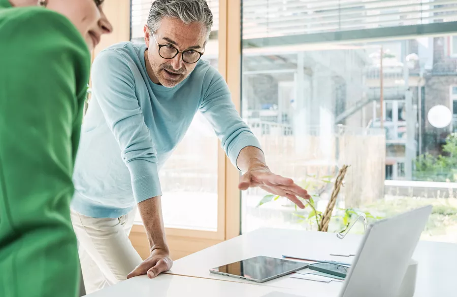 Two adults are engaged in a discussion at a bright, modern office workspace. One person gestures toward a laptop and tablet on a white desk, suggesting a collaborative or planning activity. Large windows provide natural light, and the setting appears professional and contemporary. No visible text or numeric values are present in the image.