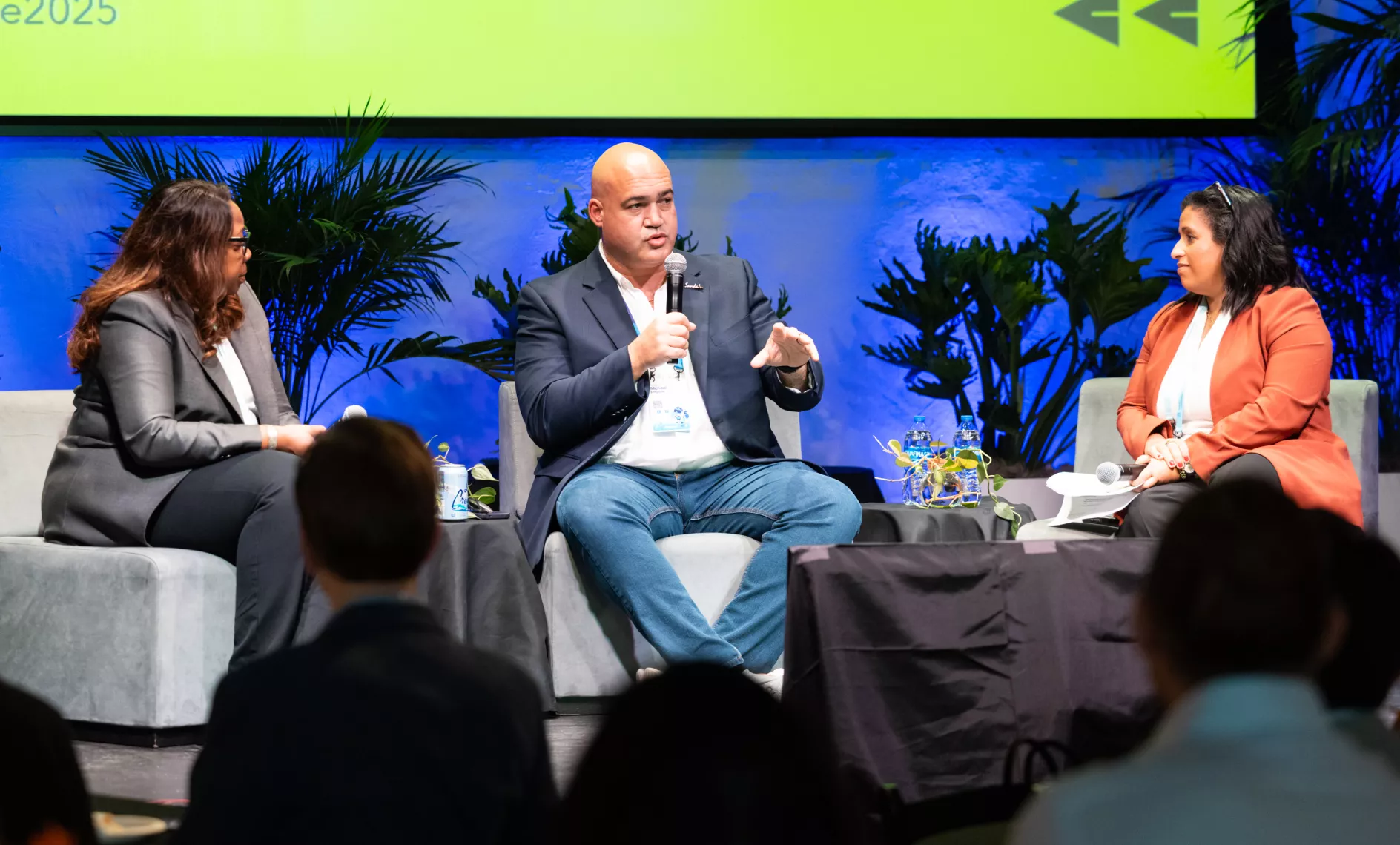 Three business professionals sit on a stage engaged in a panel discussion. The central speaker holds a microphone while addressing the audience, with two other panelists seated on either side. Attendees are visible in the foreground listening to the conversation. The backdrop features plants and a large illuminated screen, creating a formal conference atmosphere.
