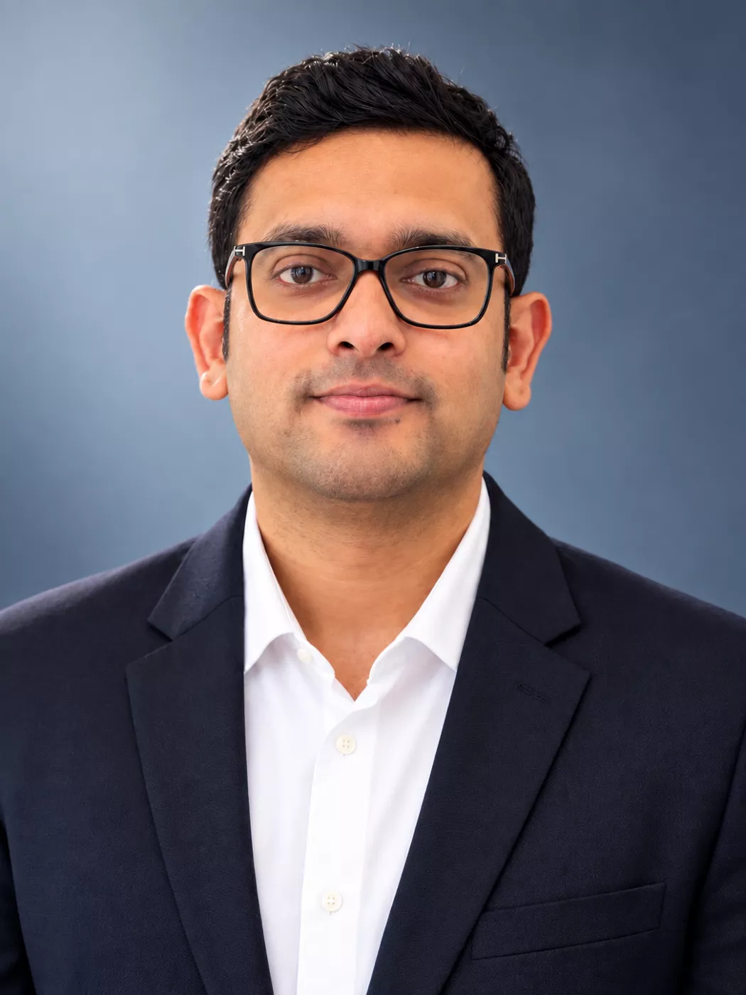Portrait of a professional man wearing a dark suit jacket and white shirt. He is facing the camera with a neutral, confident expression. The background is a smooth blue gradient, giving the image a clean corporate look. The composition is centered and well-lit, suitable for business or corporate profile use.