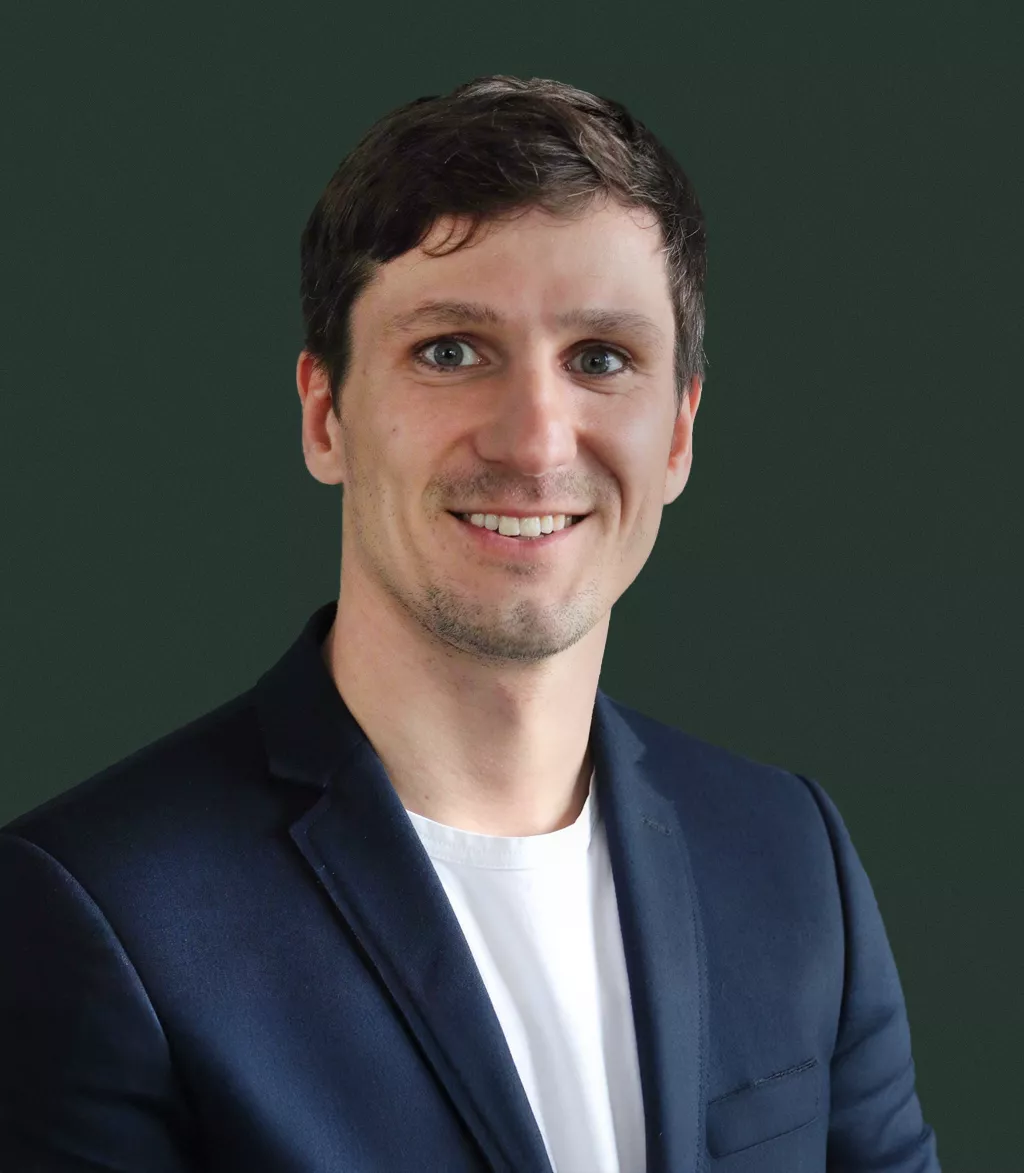 A young adult male is pictured wearing a navy blue blazer over a white crew-neck shirt. The subject is posed in front of a plain, dark green backdrop, creating a professional and minimalistic portrait style. The overall mood is formal yet approachable, with a clean and modern visual aesthetic.