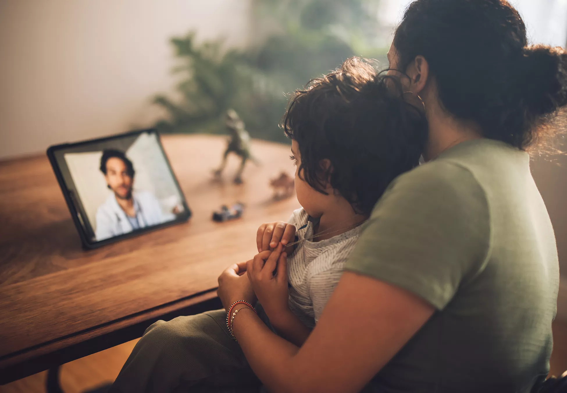 A mother sits at a wooden table holding her young child while they participate in a video call on a tablet. The tablet, propped up on the table, shows a man speaking to them from the screen. Soft natural light and indoor plants in the background create a warm, cozy atmosphere. A toy dinosaur and small cars on the table suggest a family home setting. The scene conveys connection, parenting, and remote communication.