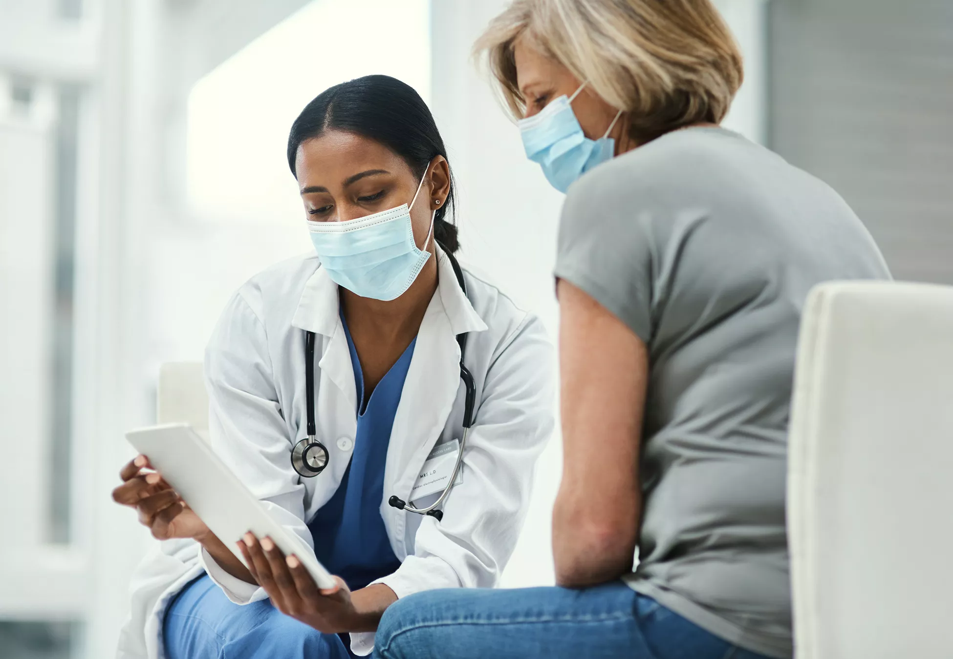 A healthcare professional wearing a white coat and stethoscope consults with an older patient. Both individuals are seated and wearing medical face masks in a bright clinical setting. The doctor holds a digital tablet, showing information to the patient and explaining details. The scene emphasizes modern healthcare, patient communication, and infection prevention.