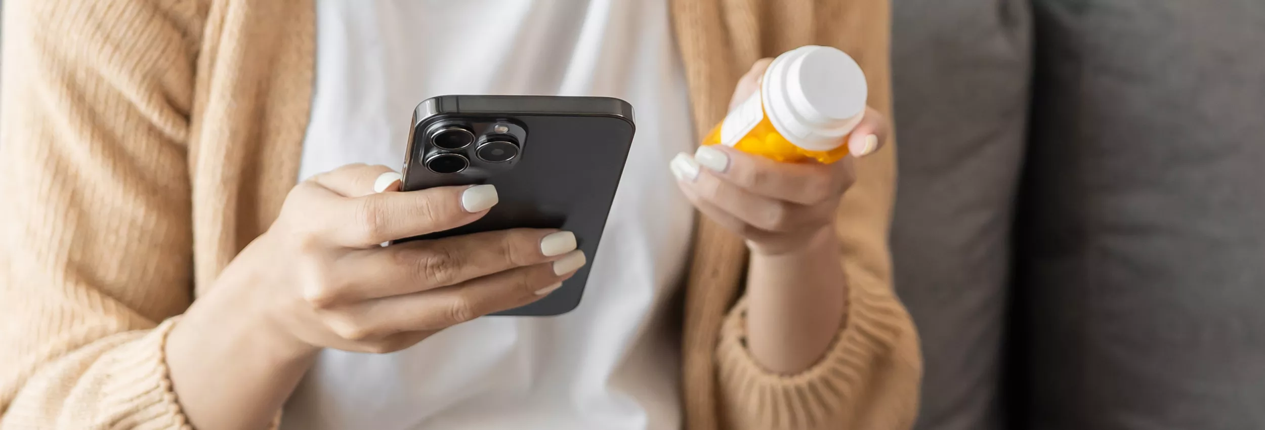 A person sits indoors holding a smartphone in one hand and a prescription pill bottle in the other. The focus is on their hands and the objects, with the rest of the body slightly out of frame. The scene suggests someone researching medication information or managing prescriptions using a mobile device. Soft, neutral tones and casual clothing create a relaxed, everyday atmosphere.