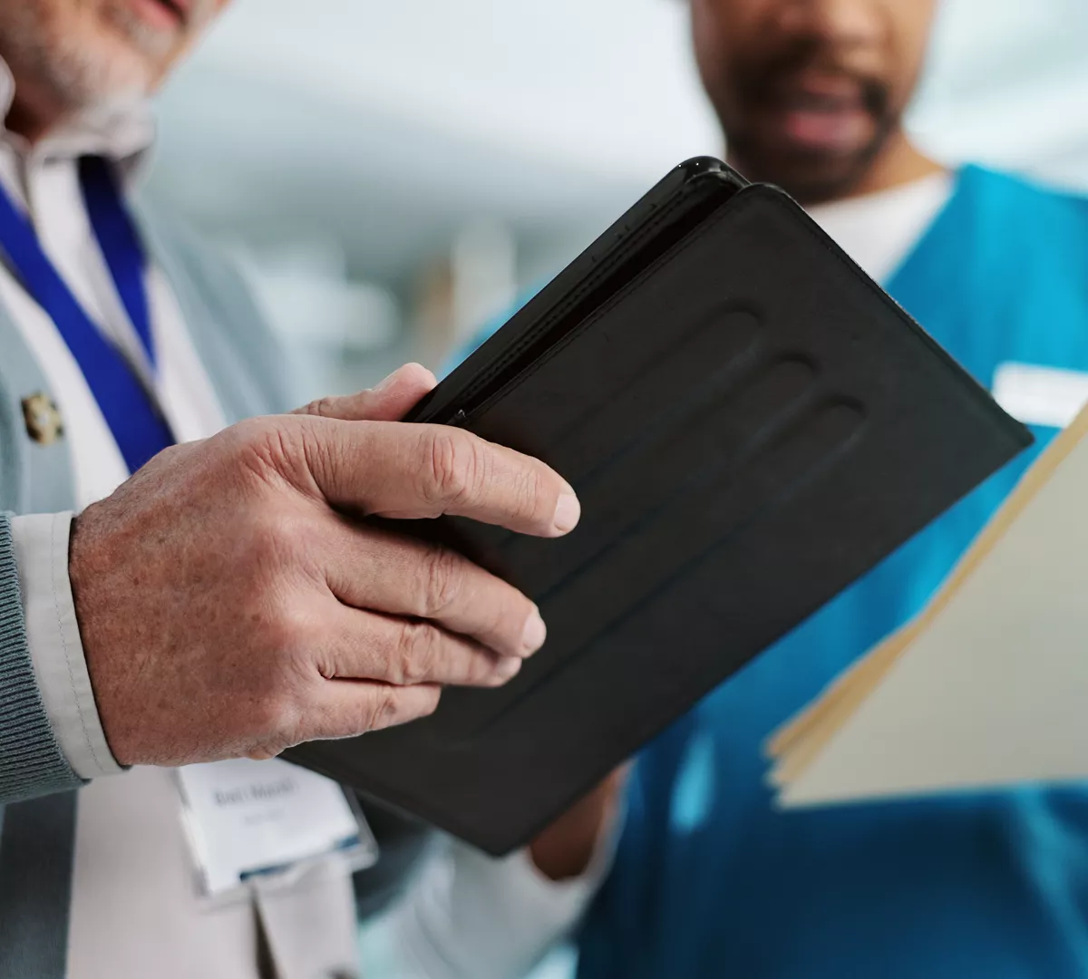 Two people stand together closely examining a tablet device. One person wears a medical scrub top and holds a folder, suggesting a healthcare or clinical setting. The other person wears a lanyard with an ID badge, indicating a professional environment. The focus is on their hands and the tablet, emphasizing digital collaboration and information sharing.