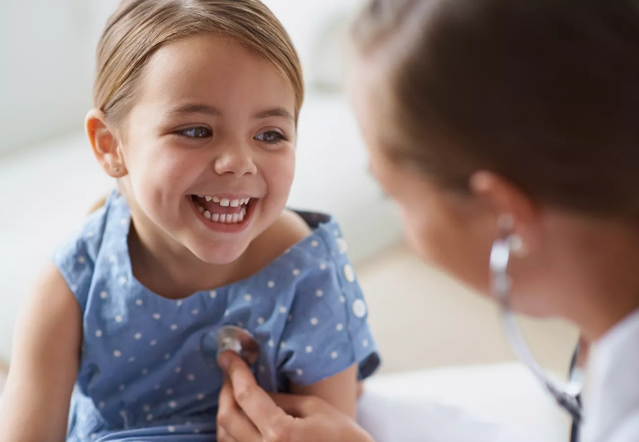 A young girl smiles brightly while a healthcare professional listens to her chest with a stethoscope. The child is wearing a blue dress with a subtle pattern in a clinical setting. The focus is on the warm interaction between the child and the medical provider. Soft lighting and close-up framing emphasize comfort, care, and trust in a healthcare environment.