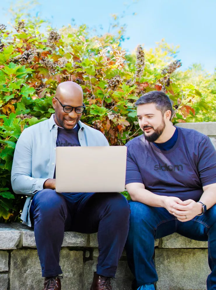 Two men sitting on a bench outside looking at a laptop 