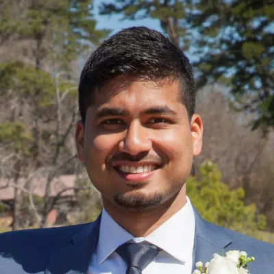 A man in a blue suit and white dress shirt smiles warmly at the camera. He is wearing a boutonniere, suggesting a formal occasion such as a wedding or celebration. The background shows trees and an outdoor setting under clear daylight. The overall mood is cheerful and welcoming.