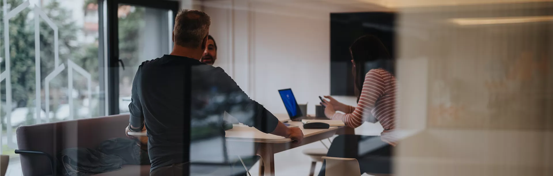 Three adults are gathered around a table in a contemporary office setting, engaged in discussion. The scene is viewed through glass, creating a reflective, dynamic visual effect. One person uses a laptop while another holds a smartphone, suggesting a tech-driven work environment. The mood is professional and collaborative, with natural light filtering in.