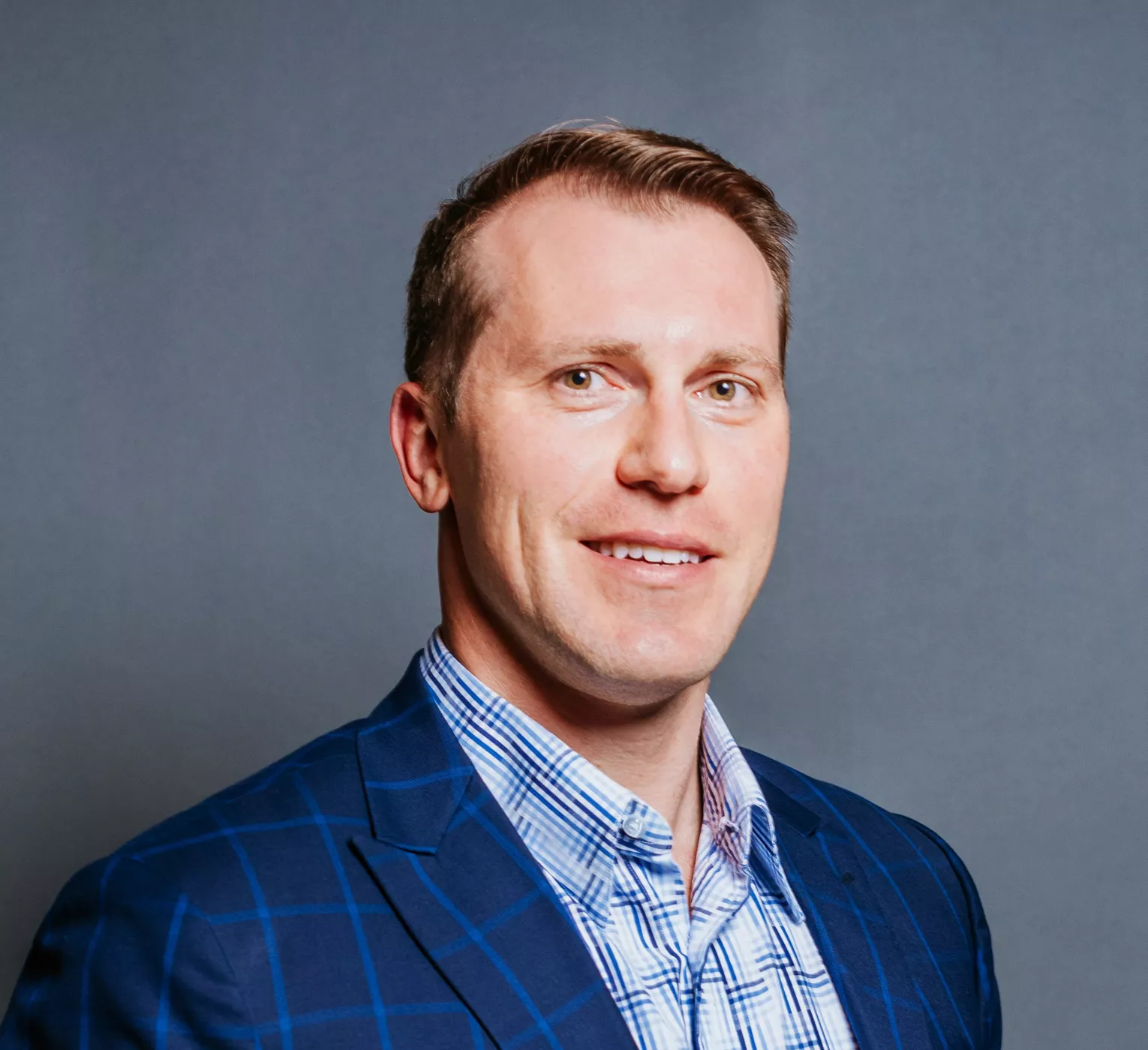 Portrait of an adult man wearing a blue checked blazer and light shirt. He is posed against a plain gray studio background, looking slightly to the side. The lighting is bright and even, highlighting his facial features and business attire. The overall image conveys a professional, corporate headshot style.