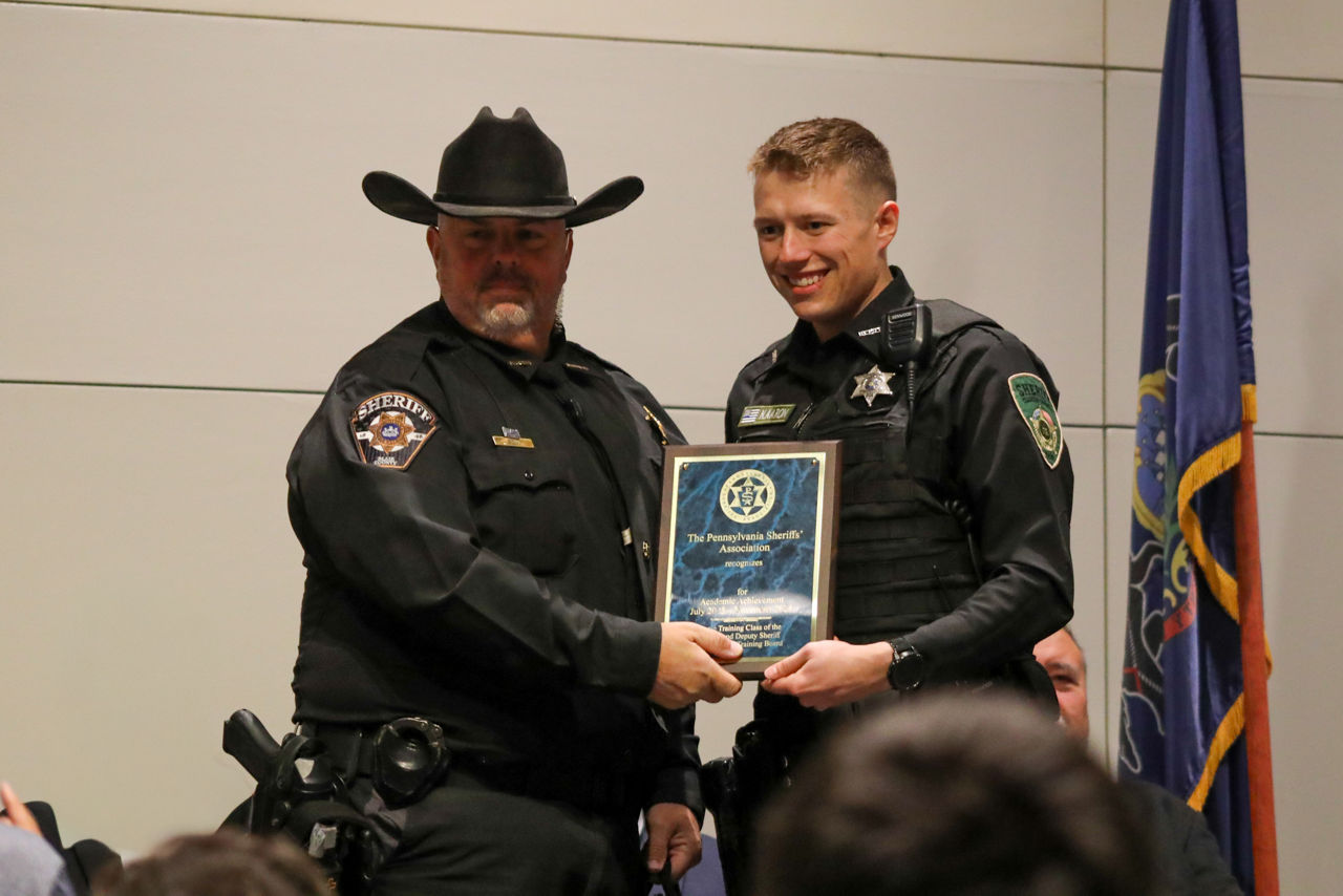 Blair County Sheriff James Ott presents the Academic Achievement Award to Clarion County Deputy Nicholas Aaron during the 72nd Sheriffs’ & Deputy Sheriffs’ Basic Training Academy Commencement Ceremony. Deputy Aaron smiles as he accepts the plaque recognizing his accomplishment.