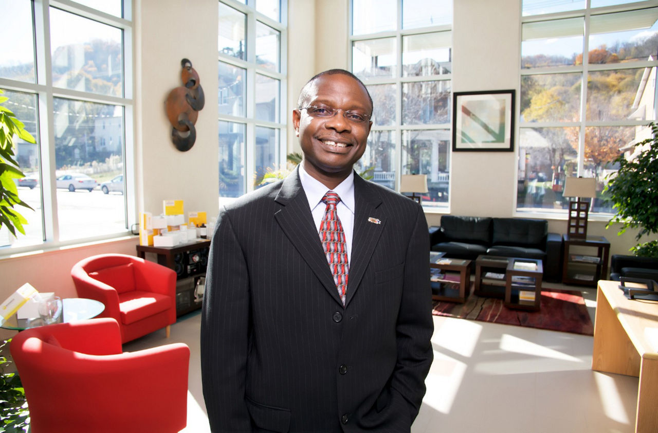 A man wearing a suit smiles for a picture in a brightly lit office lobby with many windows and modern furniture