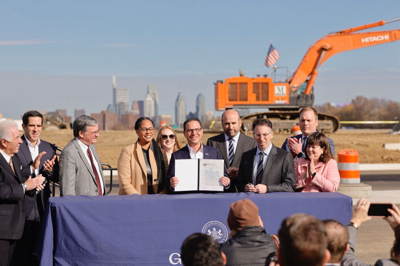 Governor Josh Shapiro, surrounded by a group of government officials and business leaders, smiles and shows an exectutive order document for the just-announced Permit Fast Track program. The Philadelphia skyline and construction equipment are visible in the distance.