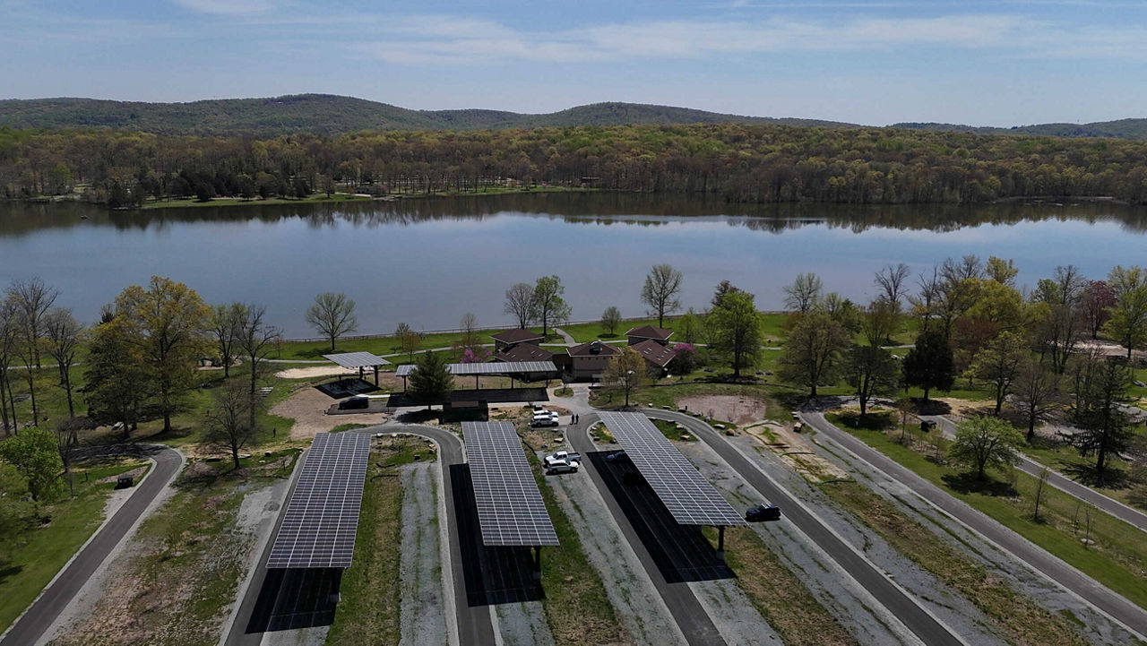 An aerial view of a serene lake surrounded by green hills and trees in the background. In the foreground, several solar panel arrays are installed in a parking area, with a building visible nearby.