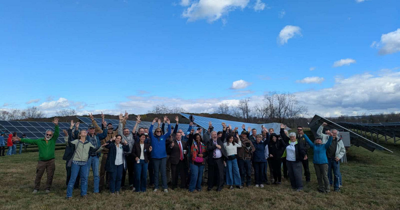 A large group of people stands outdoors in front of solar panels, all raising their arms in celebration. 
