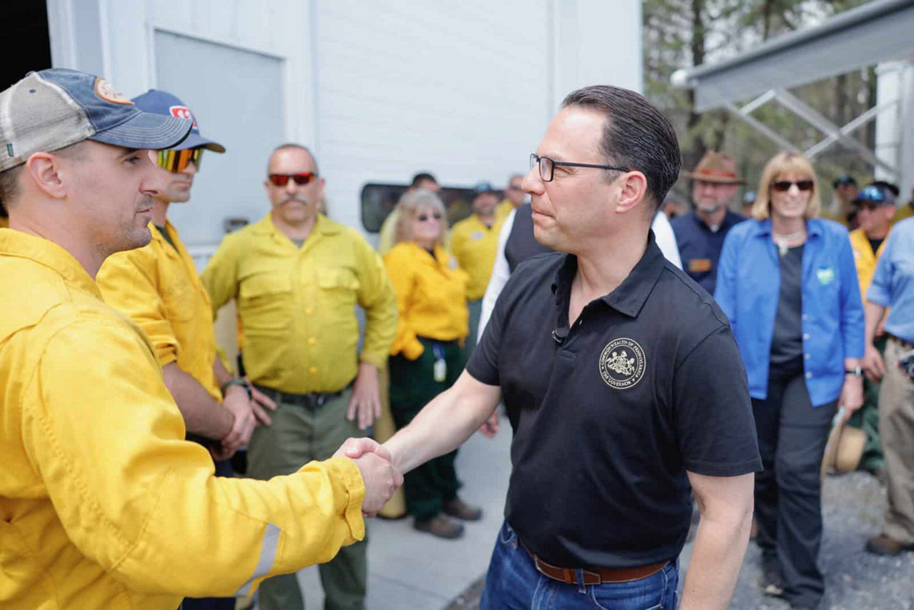 Pennsylvania Governor Josh Shapiro, wearing a blue shirt, shakes hands with a member of a group in yellow firefighting uniforms outside a building. 